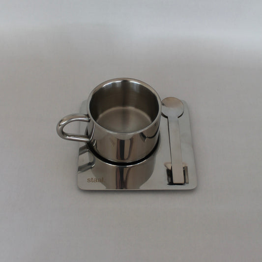 En stål kop med hank placeret på en firkantet stålunderkop, der har en lille ske ved siden af. Designet er minimalistisk med en glat, poleret overflade og logoet 'staal.' synligt på underkoppen, præsenteret mod en neutral baggrund.