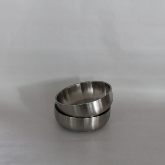 To metal skåle i et minimalistisk design, hvor den ene skål er stablet delvist ovenpå den anden. Skålene har en glat, poleret overflade og præsenteres mod en neutral baggrund.