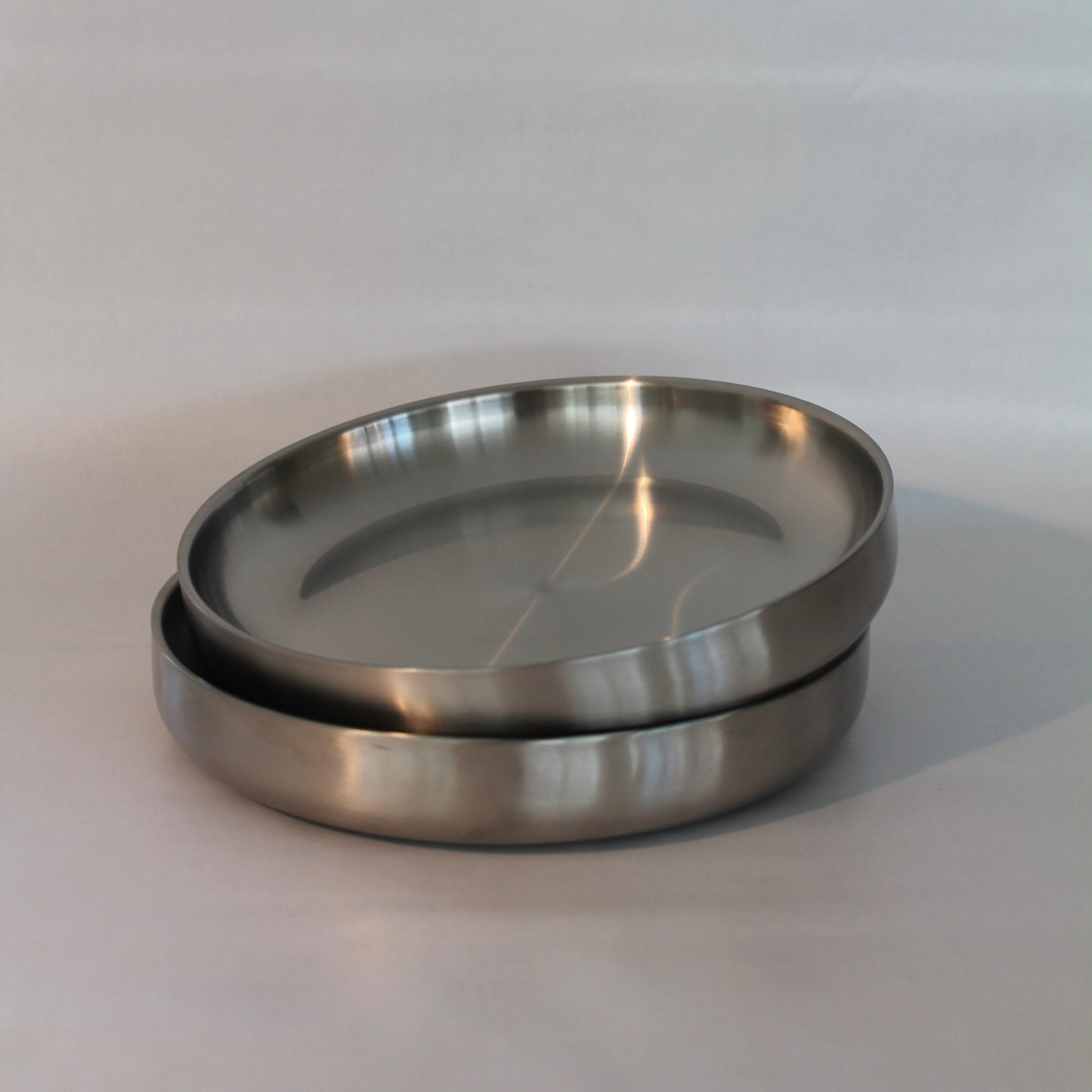 To metal tallerkener stablet ovenpå hinanden i et minimalistisk design med en glat og poleret overflade. Tallerkenerne er præsenteret mod en neutral baggrund.