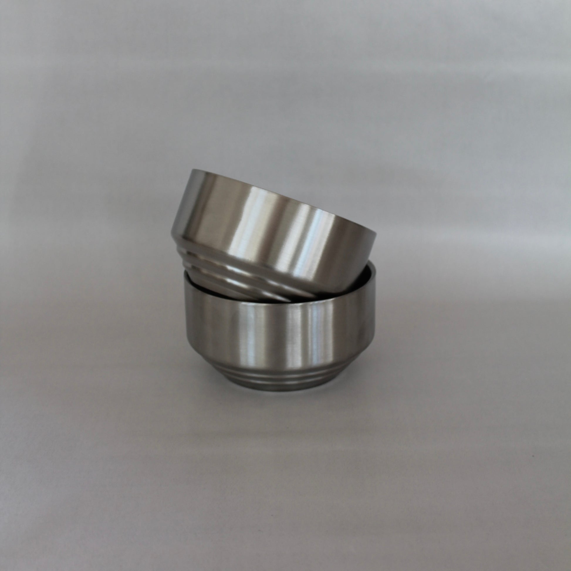 To metal skåle med rillede detaljer stablet ovenpå hinanden, hvor den øverste skål er vippet let til siden. Skålene har en glat, poleret overflade og står mod en neutral baggrund.