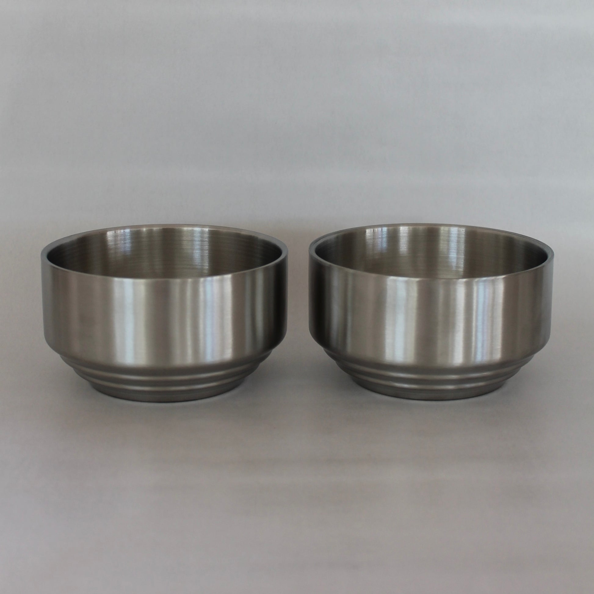 To ens metal skåle med rillede detaljer placeret side om side. Skålene har en glat, poleret overflade og står mod en neutral baggrund.