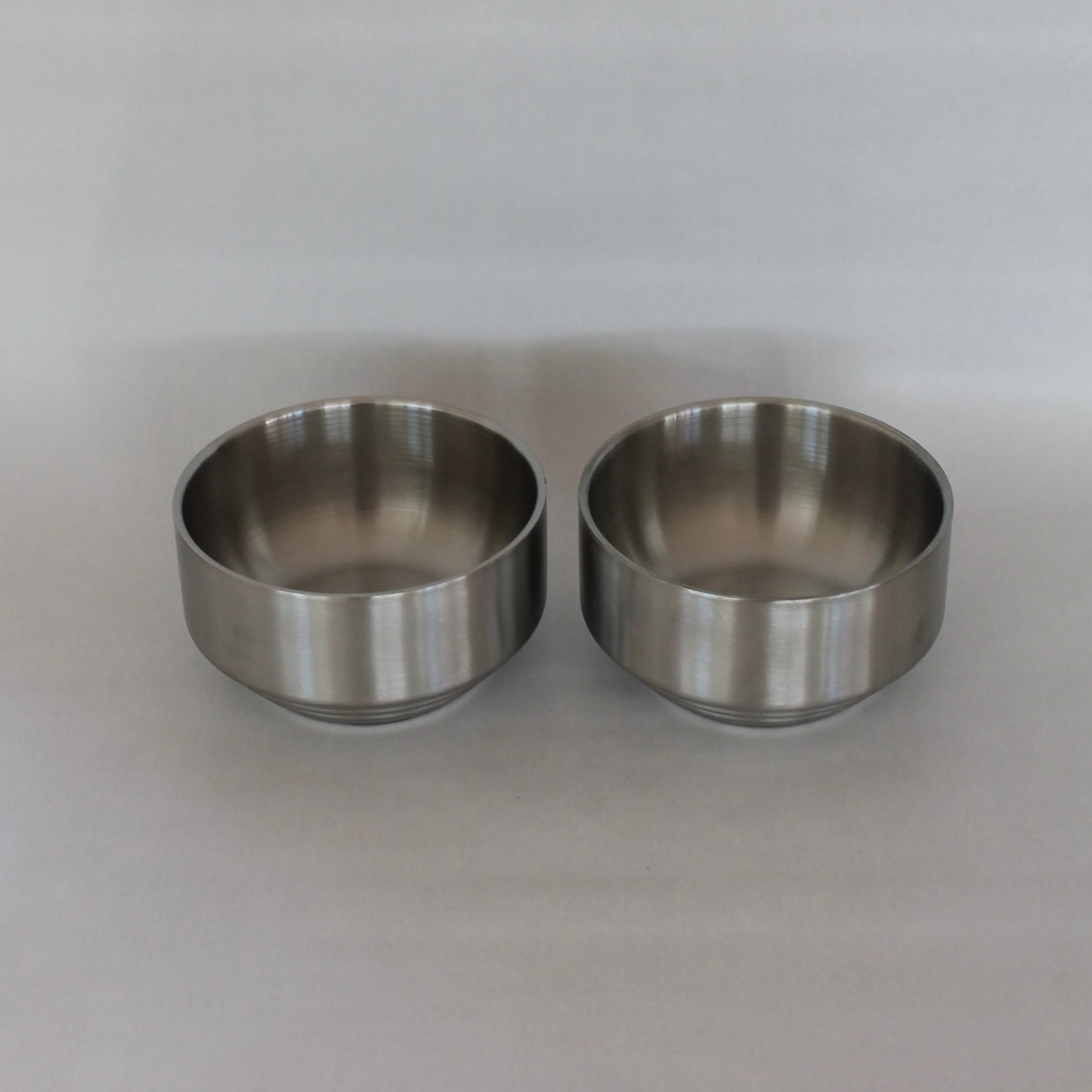To ens metal skåle med en glat og poleret overflade, placeret side om side på en neutral baggrund. Skålene har en enkel og minimalistisk form.