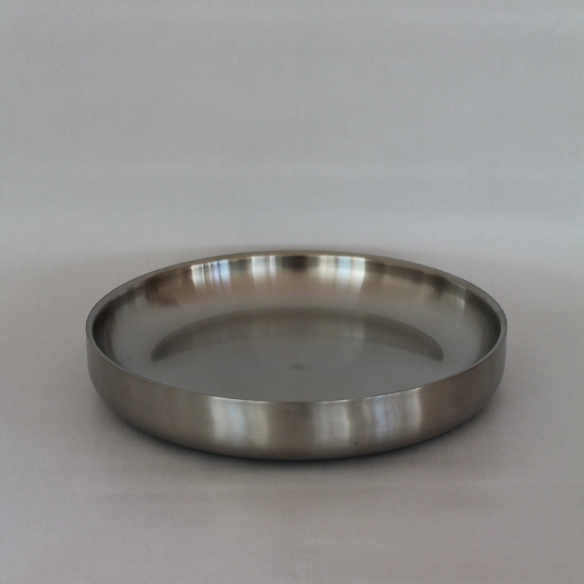 En rund, lav metalbakke eller metal tallerken med en glat og poleret overflade i en minimalistisk stil, set fra en let vinkel mod en neutral baggrund.