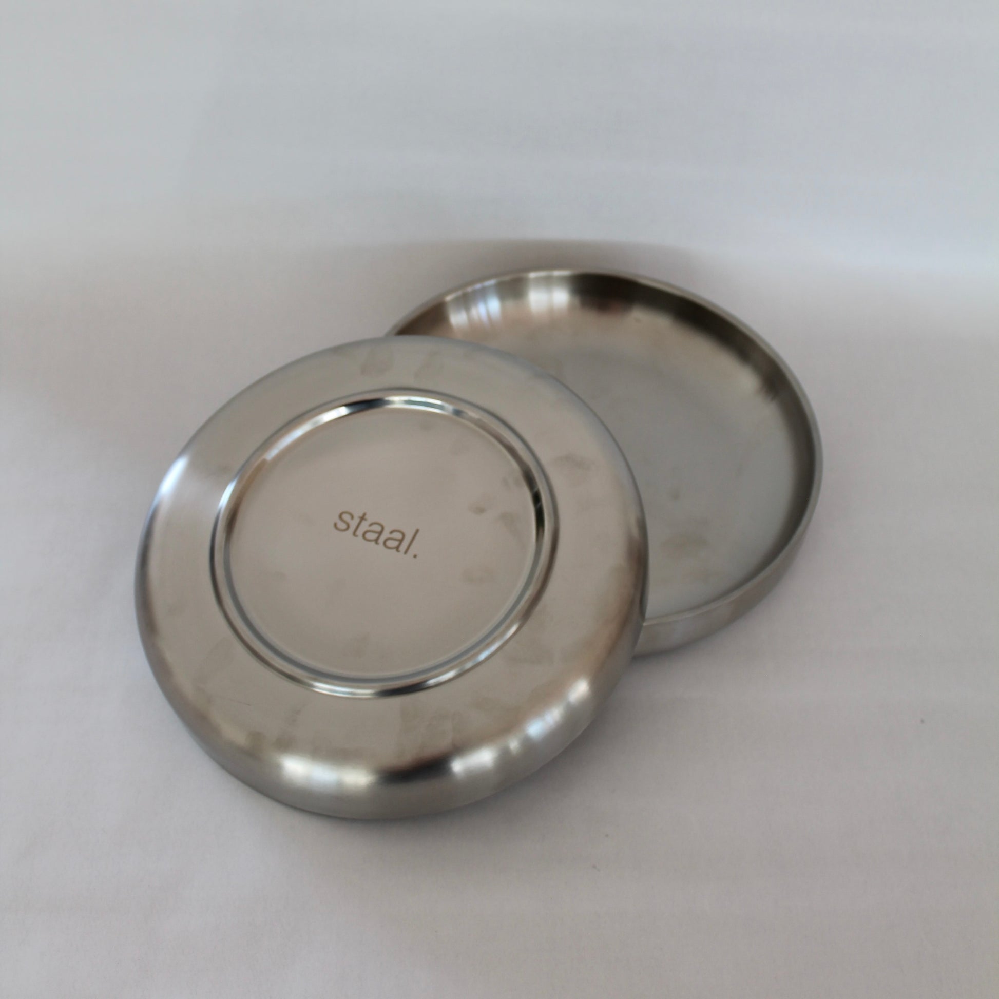 To metal tallerkener i minimalistisk design, hvor den ene tallerken ligger ovenpå den anden med bunden opad. Bunden viser et indgraveret logo med teksten 'staal.' på en neutral baggrund.